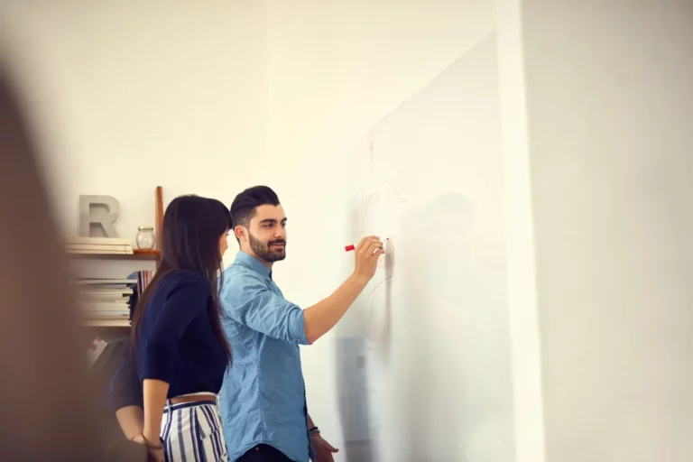 Two people writing on a whiteboard in an office.