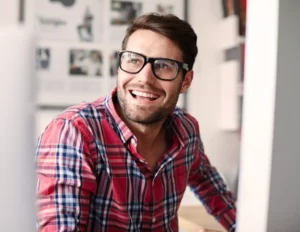 A man wearing glasses is smiling in front of a computer.