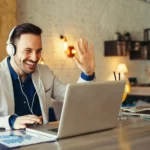 A man wearing headphones is waving at a laptop.