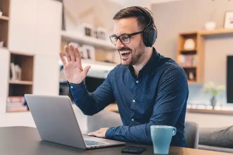 A man wearing headphones is using a laptop and waving at the camera.