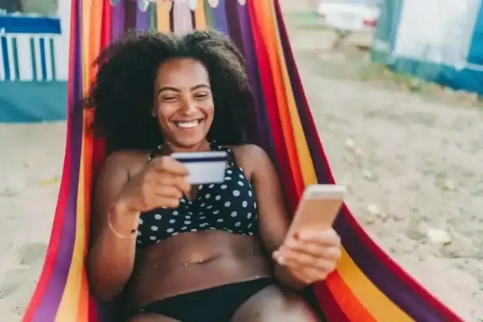 A woman in a garment sitting in a hammock holding a credit card and a phone.