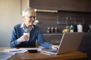 An older woman sitting at a table with a cup of coffee and a laptop.