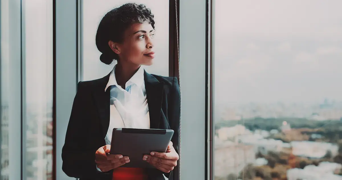 A business woman looking out of a window with a tablet.