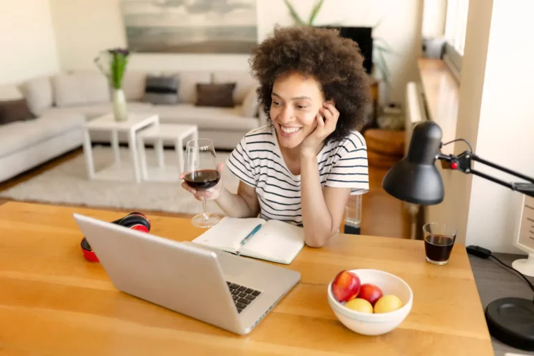 A woman is sitting at a table with a glass of wine and a laptop.