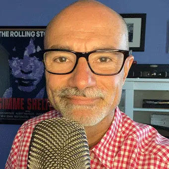 A bald man with glasses and a microphone in front of a poster.