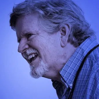 A man with a beard is laughing in front of a blue background.