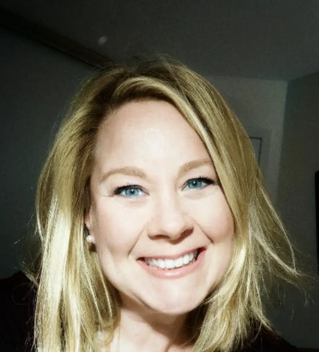 A woman with blonde hair and blue eyes smiling at the camera in a well-lit room.