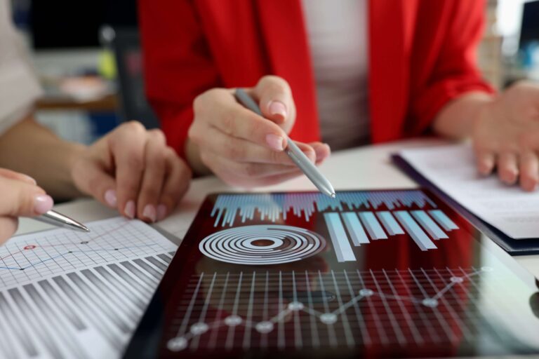 Two individuals analyze data on a tablet displaying bar and line graphs. One person points at the display with a pen, while the other holds documents with printed charts.