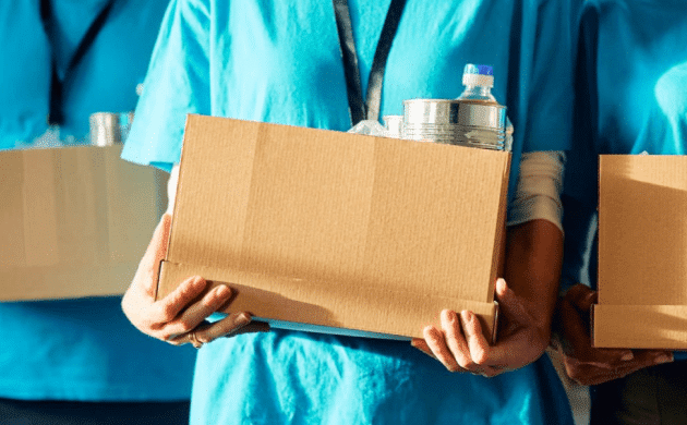 Three people wearing blue shirts hold cardboard boxes filled with canned goods and bottled water, showcasing synergy in their efforts to support those in need.