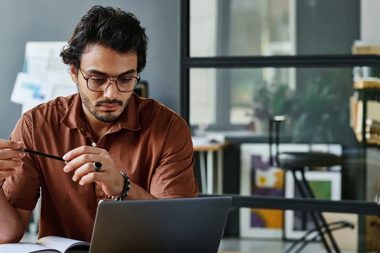 A man with glasses and a beard, wearing a brown shirt, sits at a table looking at a laptop screen, holding a pen. There are office items and a large glass window in the background.