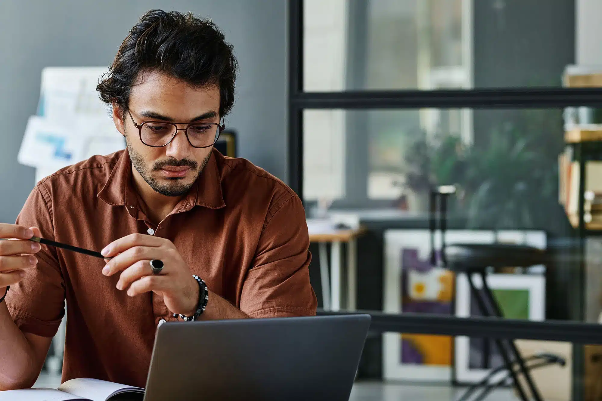 A man with glasses is seated at a desk, looking at a laptop screen while holding a pen in his hand in an office setting.