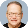 A man with short, curly gray hair, glasses, and a slight smile is wearing a black shirt. The background is a plain blue-gray color, giving the scene a calm synergy.