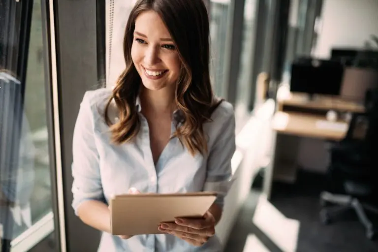 A woman stands by a window holding a tablet, smiling while looking outside, with a blurred office space visible in the background.