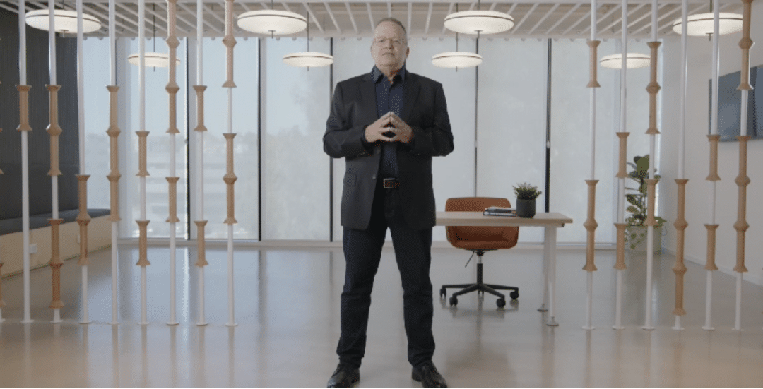 A man in a dark suit stands in an open, modern office space with large windows, a wooden desk and chair, a small plant, a book, and a notebook. Vertical wooden spindles add to the decor's synergy.