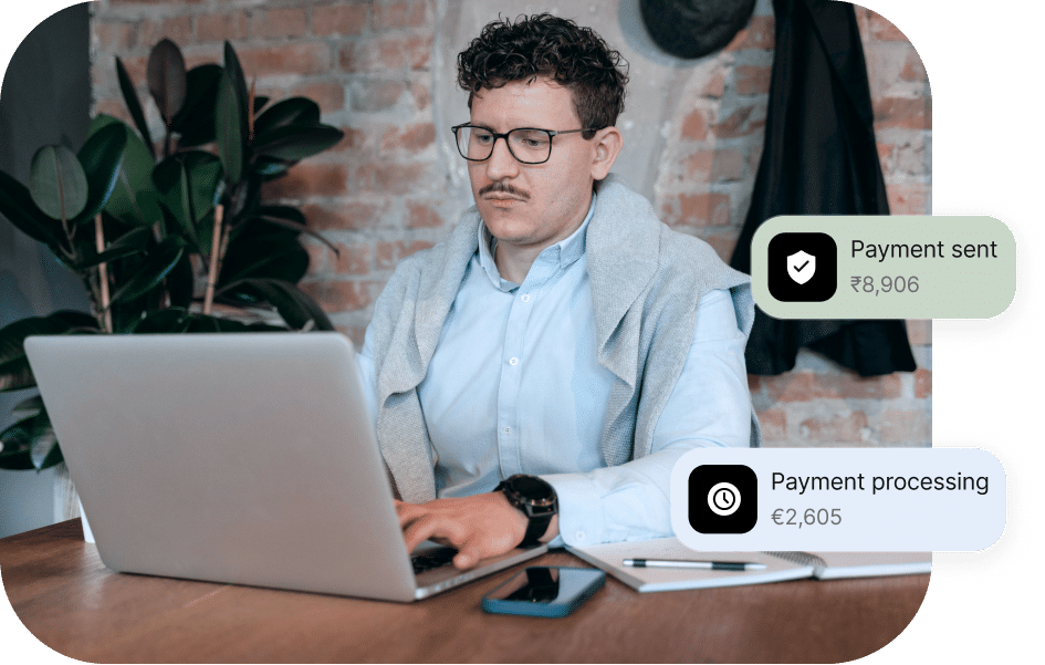 A man with curly hair and glasses works on a laptop at a wooden desk, with notifications of payment processing in different currencies.