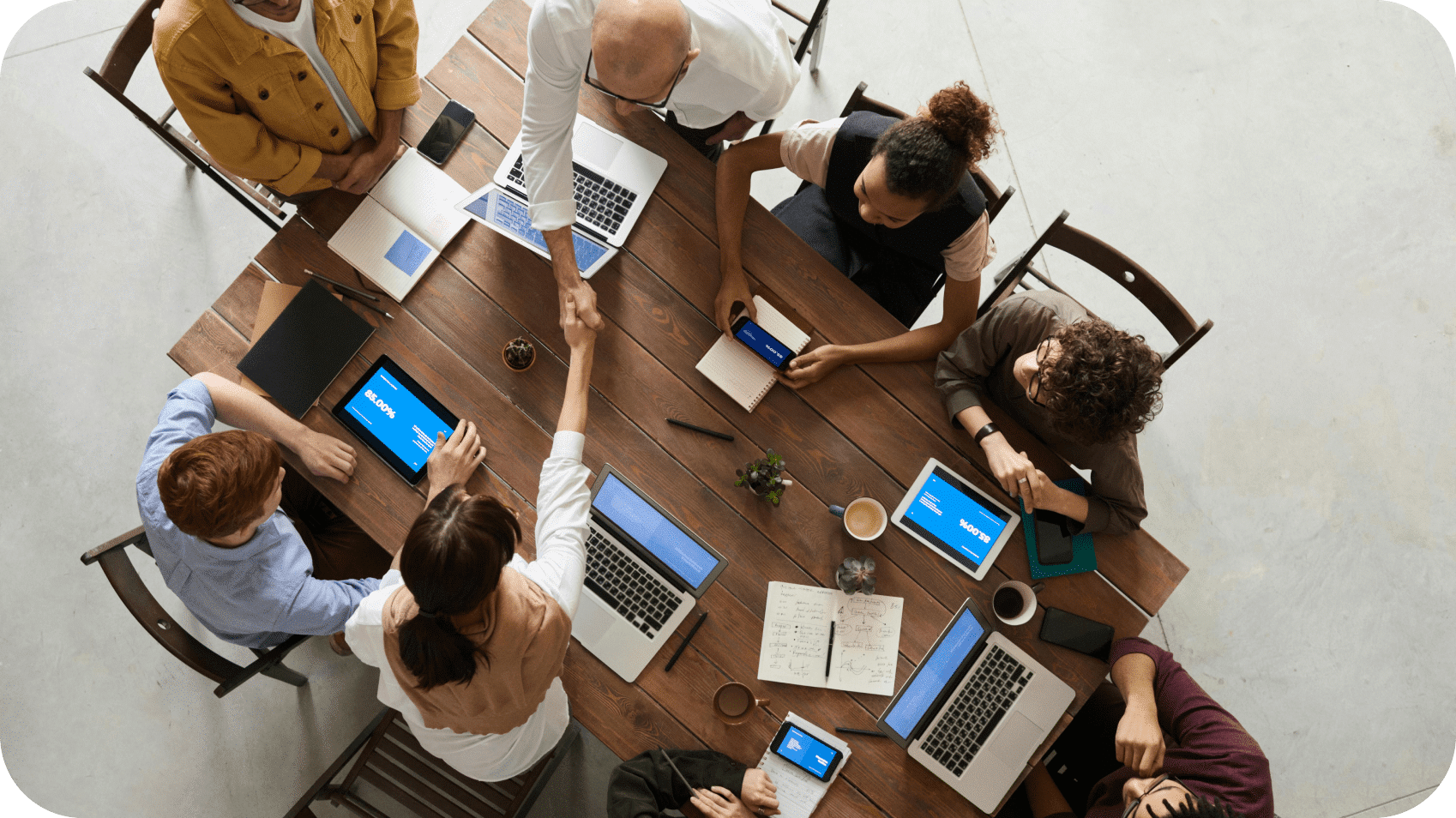 A group of eight people sitting around a wooden table with laptops and tablets, engaged in discussion about global payments. Two people are shaking hands in the center of the table, likely sealing a deal involving Tipalti's payment automation solutions.