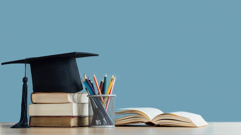 A graduation cap rests on stacked books beside an open book and a container of colored pencils, symbolizing learning that fuels various industries, all on a desk.