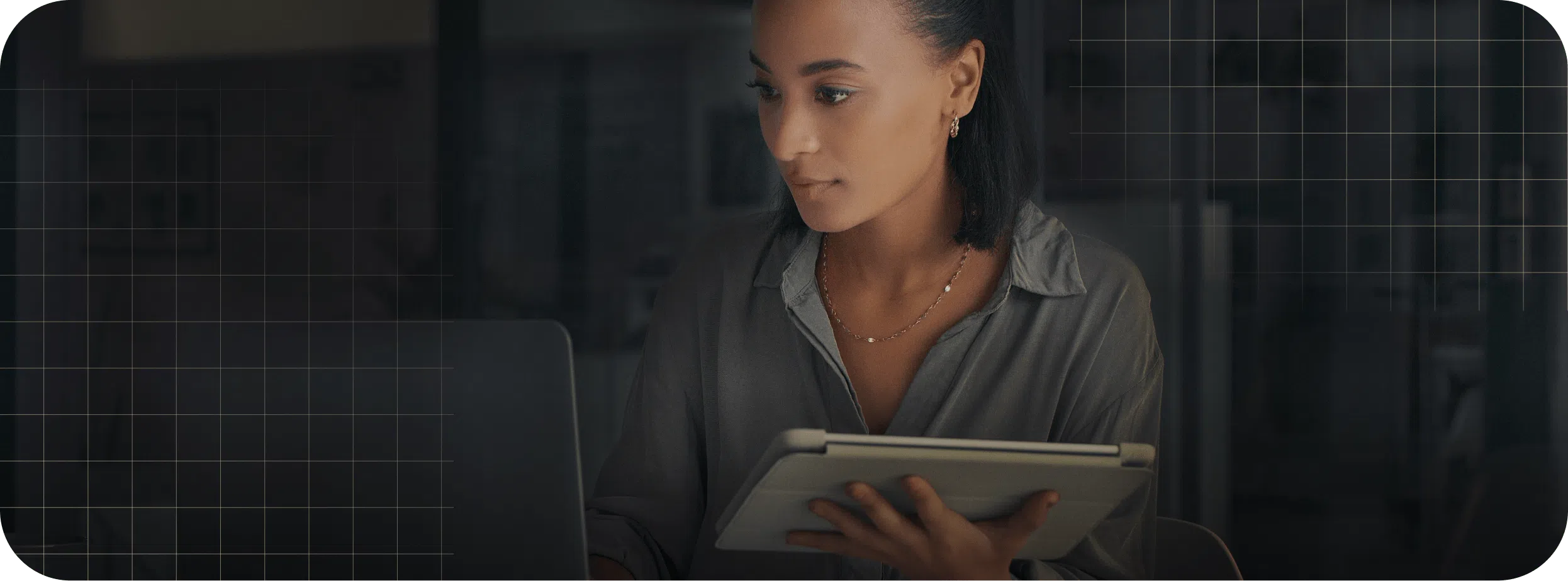 A person sits at a desk, looking at a laptop screen while holding a tablet. The background is dark with a grid pattern overlay, creating an atmosphere of focused synergy_home productivity.