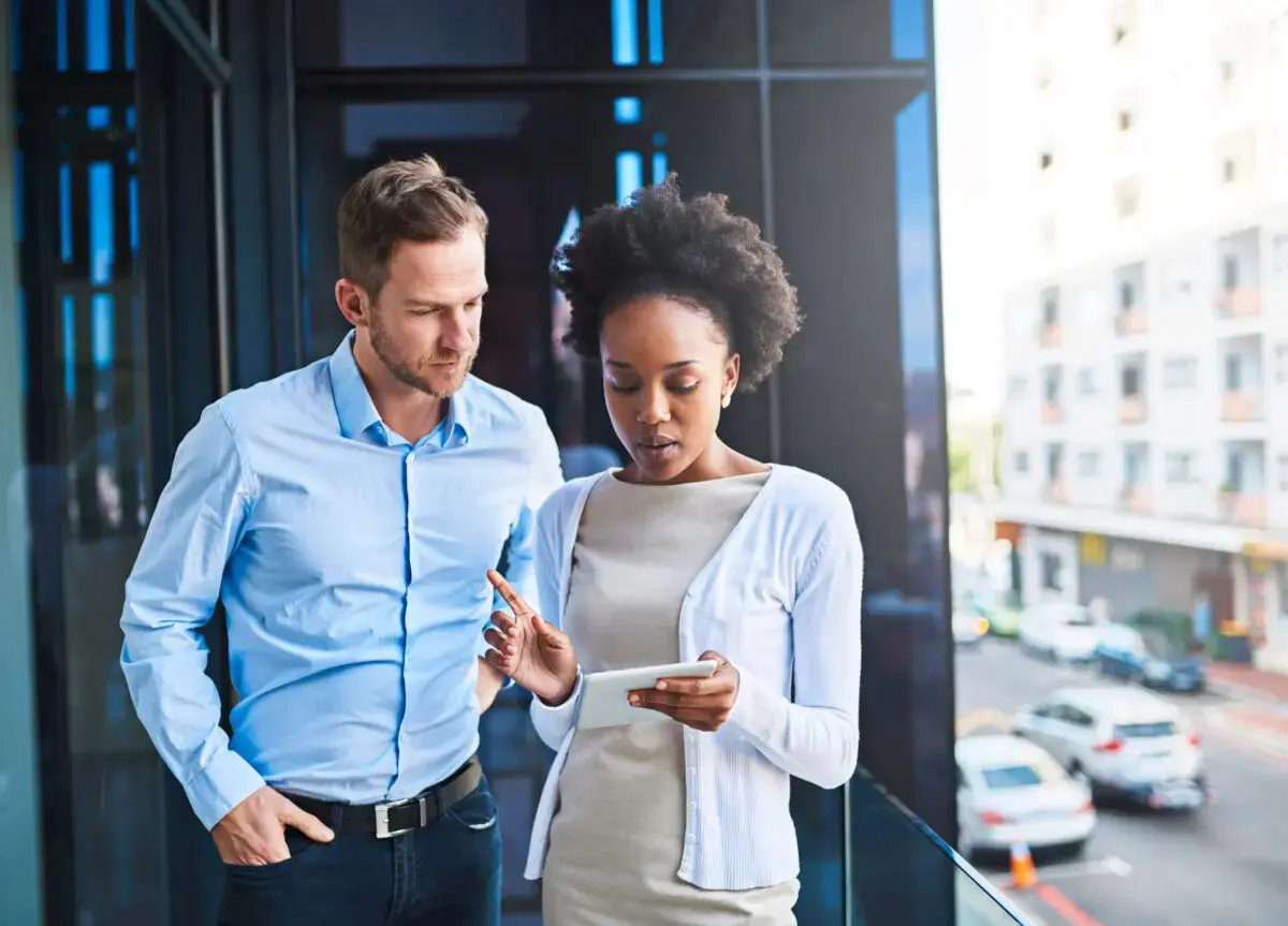 A man and woman stand on a balcony, looking at a tablet together. In the urban setting behind them, they discuss how their VAT number plays a crucial role in their business endeavors.