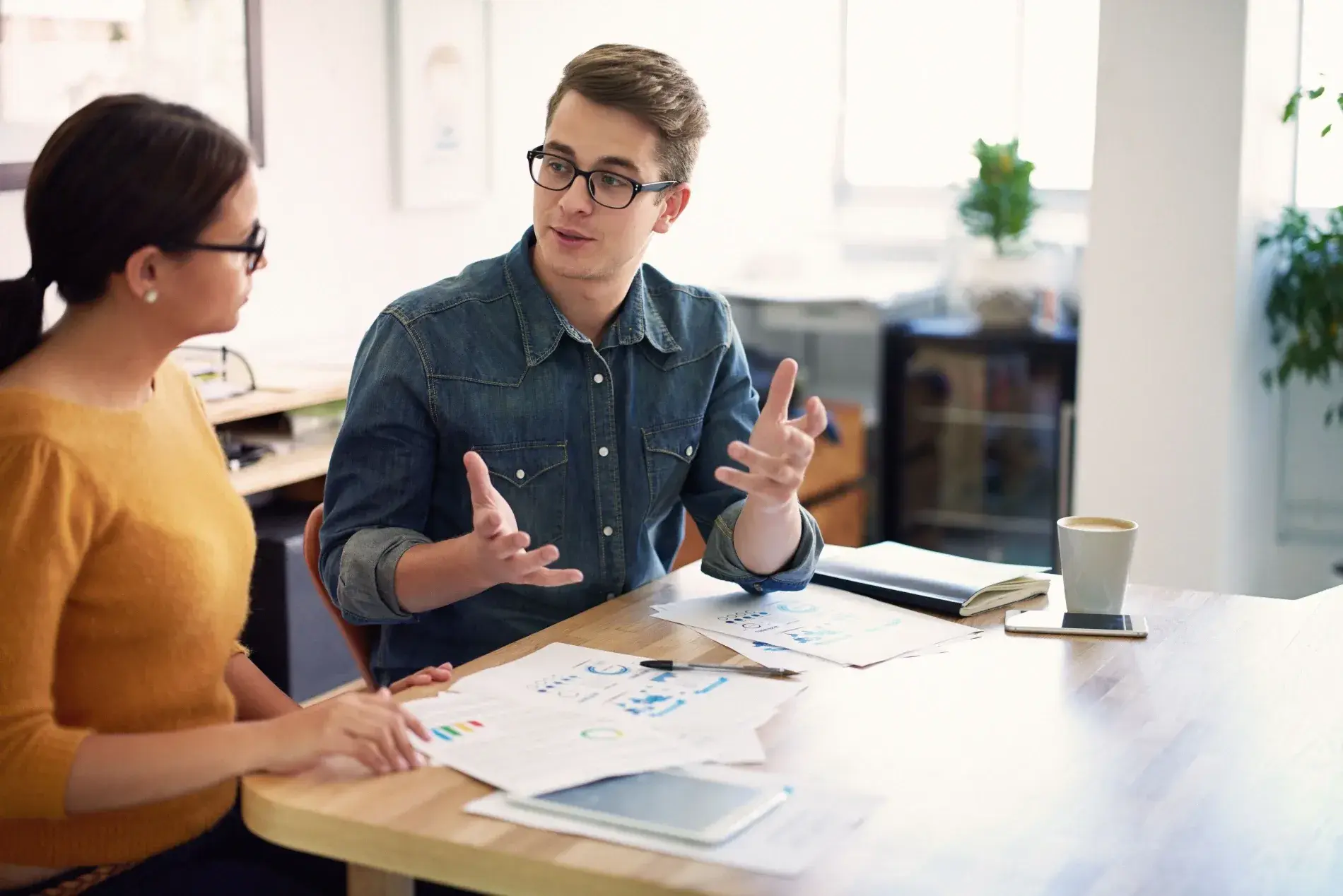 Two people are sitting at a table, discussing documents and a tablet in front of them. One gestures passionately about the implications of VAT on digital services while a cup of coffee rests nearby, undisturbed by their intense conversation.