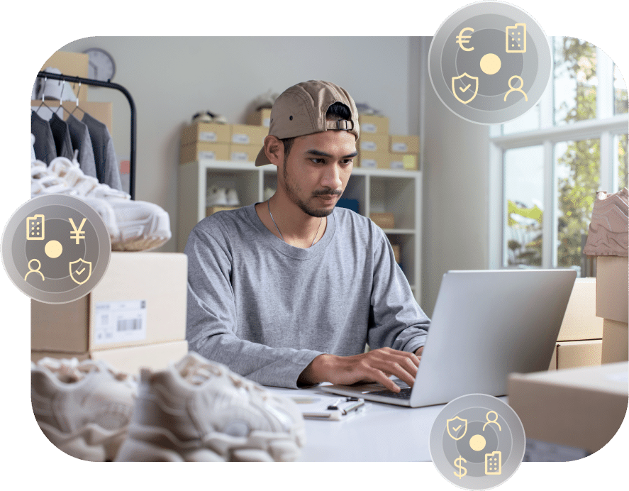 A man wearing a cap and grey shirt works on a laptop surrounded by shoe boxes. Three circular icons depicting business-related graphics are overlaid on the image.