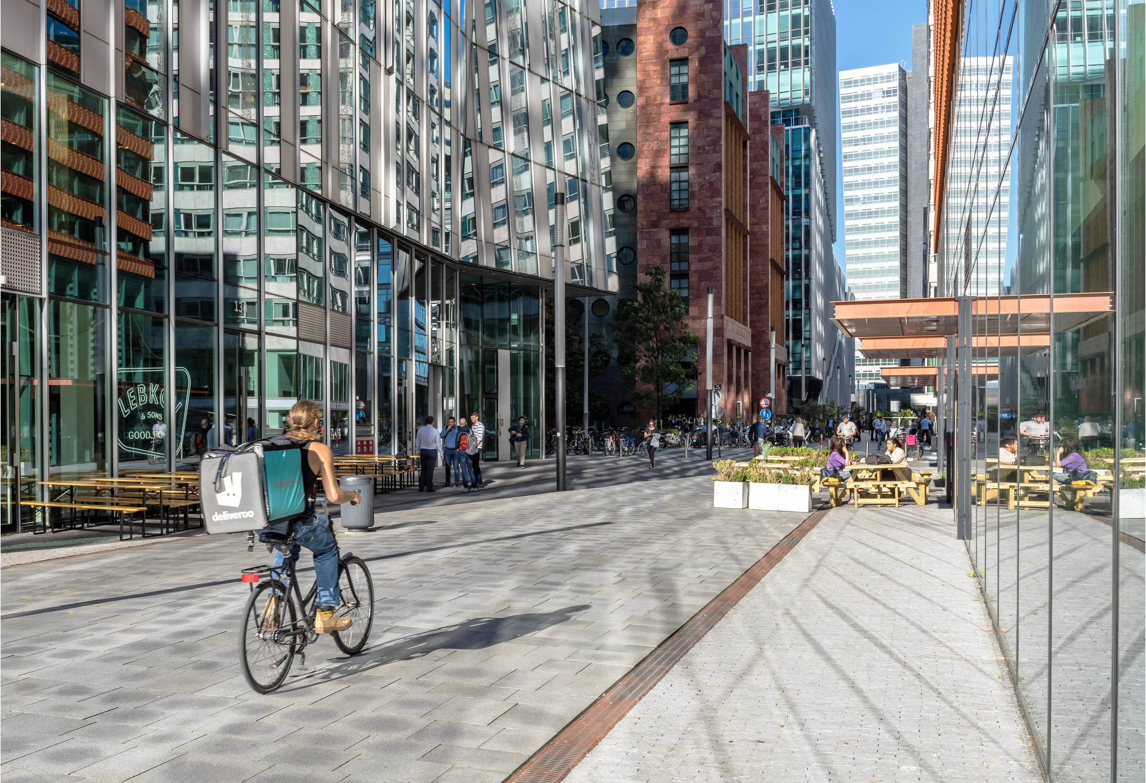 Urban street scene with modern glass buildings, a cyclist carrying a delivery bag, pedestrians, and outdoor seating area on a sunny day.