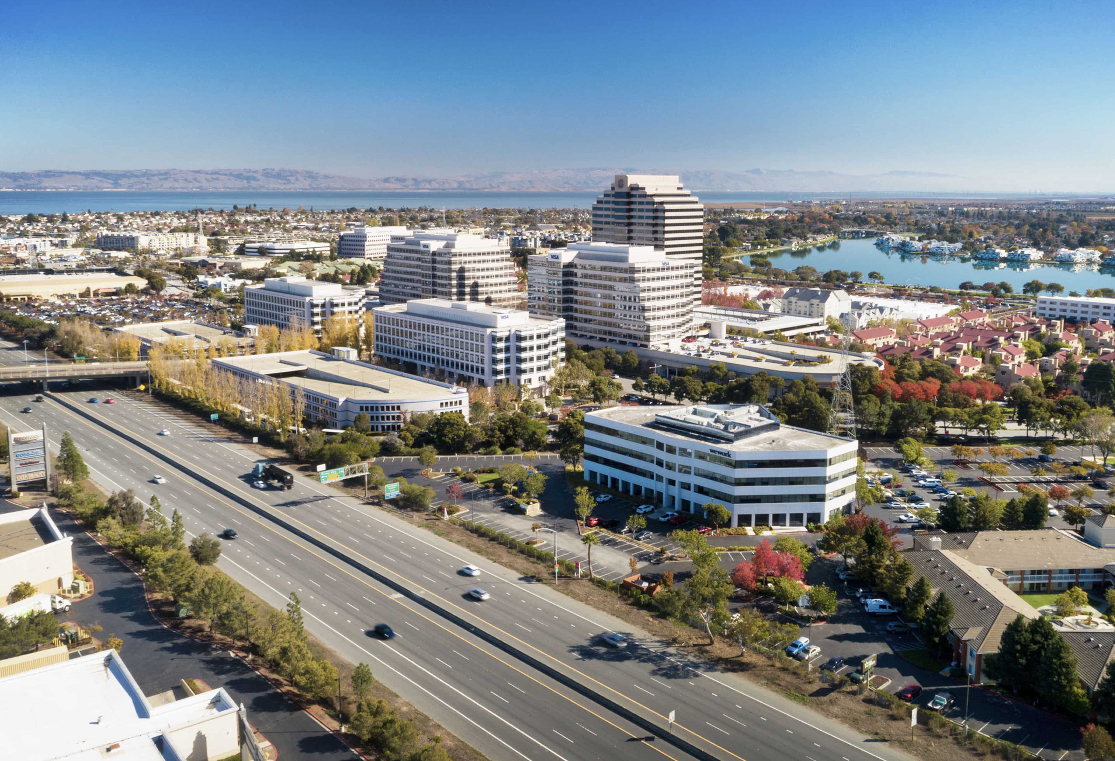 Aerial view of an urban area featuring office buildings, a highway with vehicles, and a residential neighborhood with water bodies in the background under a clear blue sky.