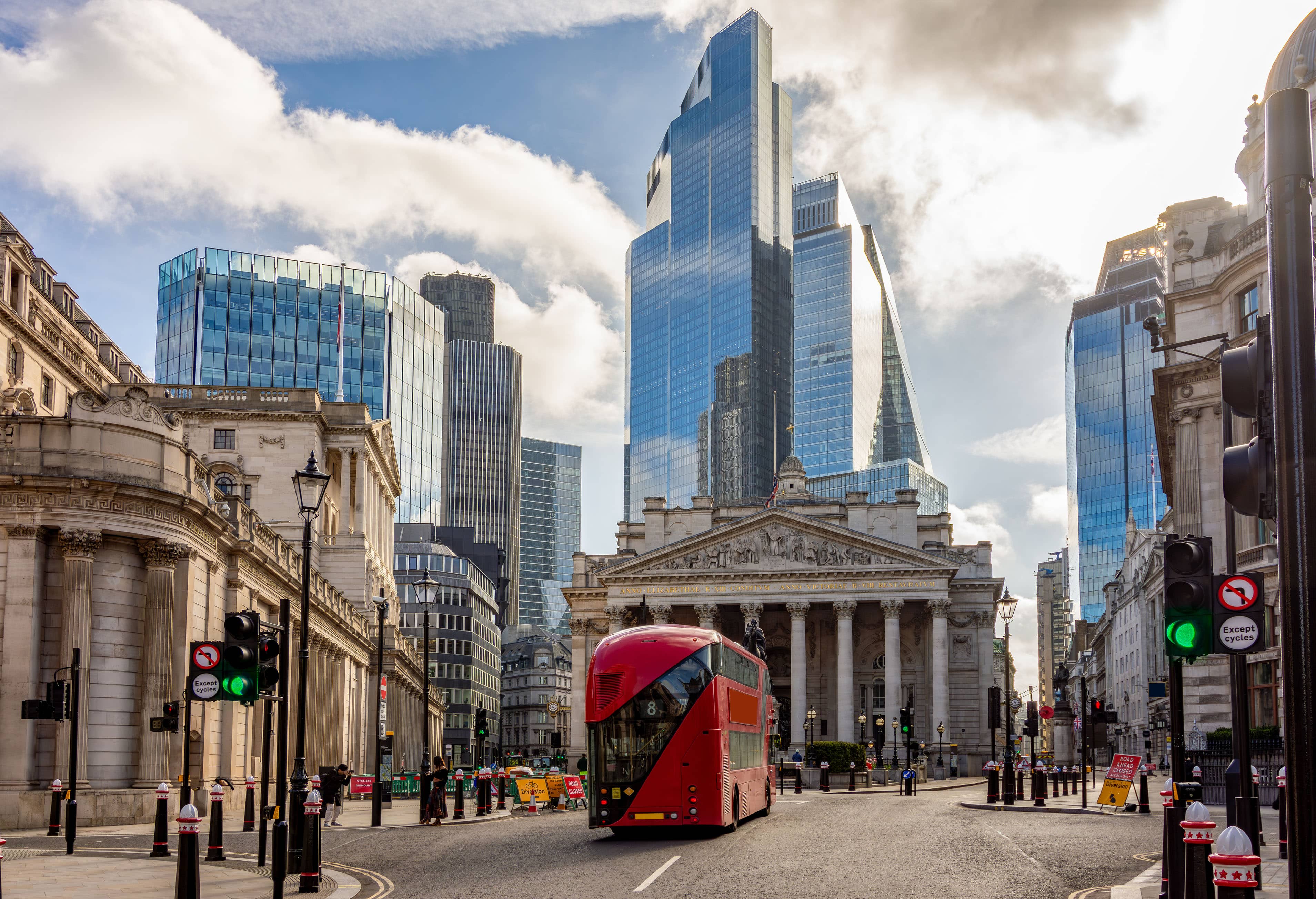 Red double-decker bus on a street in a city with modern skyscrapers and classical architecture under a blue sky with clouds.