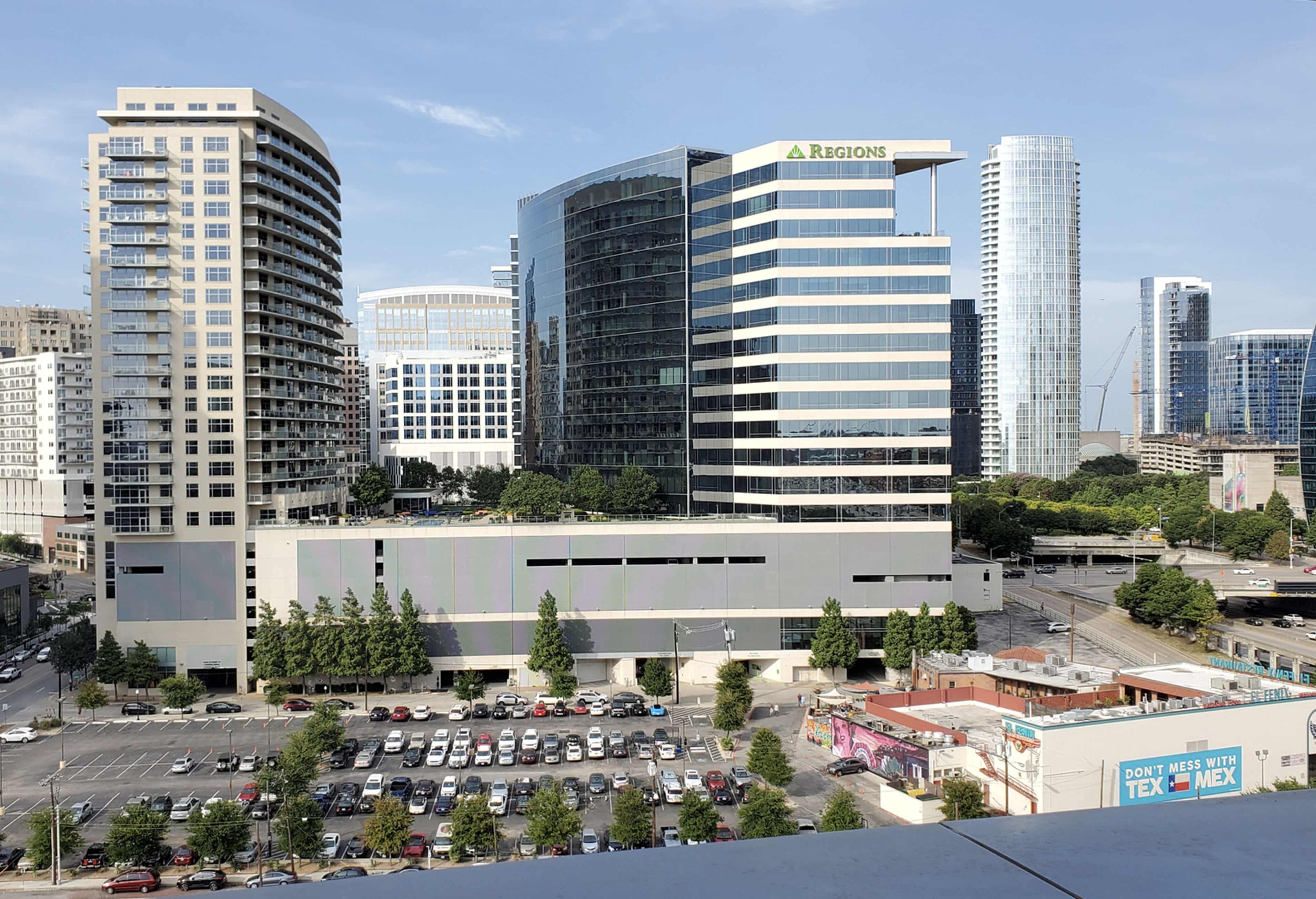 Cityscape showing a cluster of modern high-rise office and residential buildings with a parking lot and a street in the foreground under a clear sky.