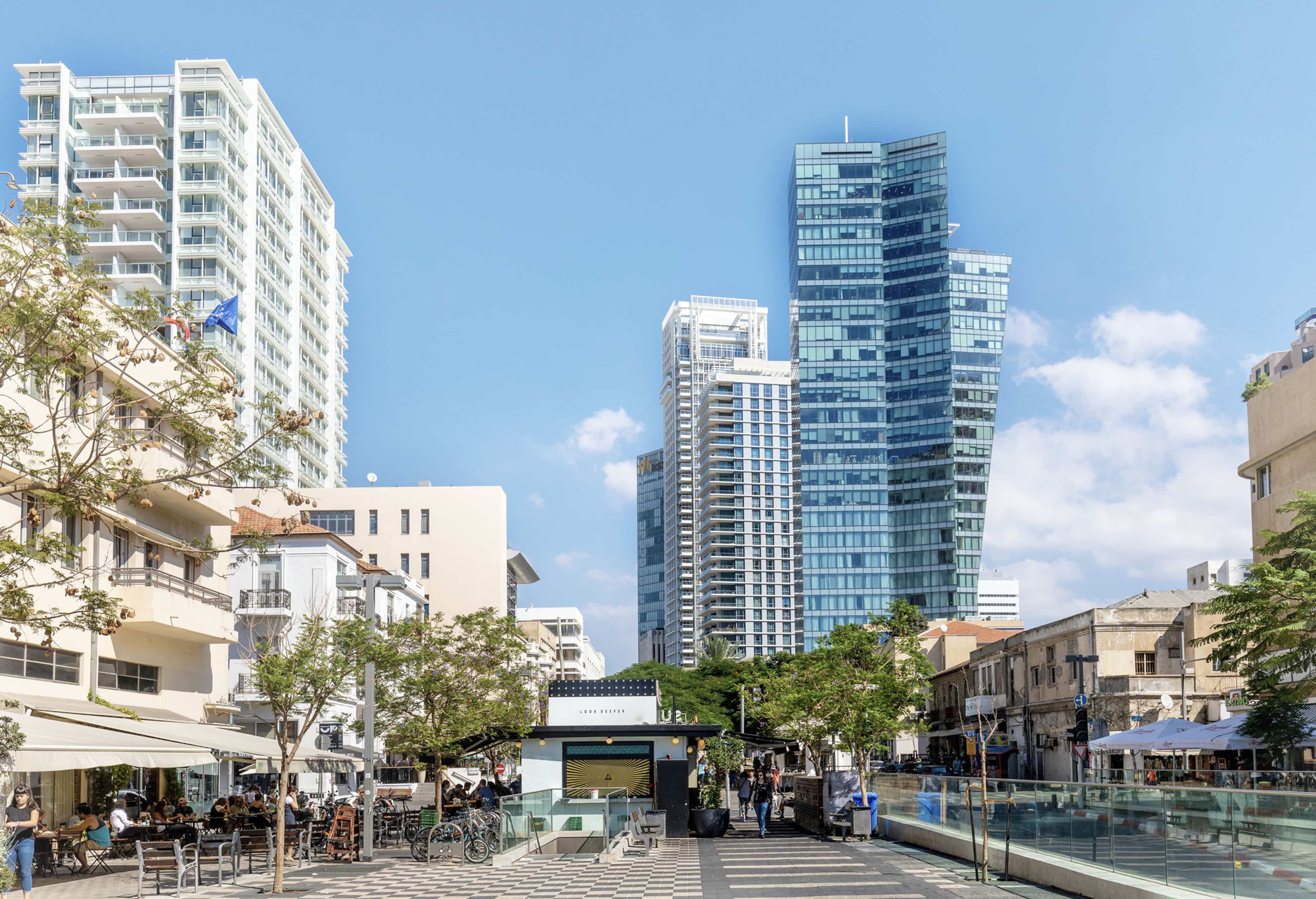 A city street with modern high-rise buildings, outdoor cafes, and pedestrians under a clear blue sky.