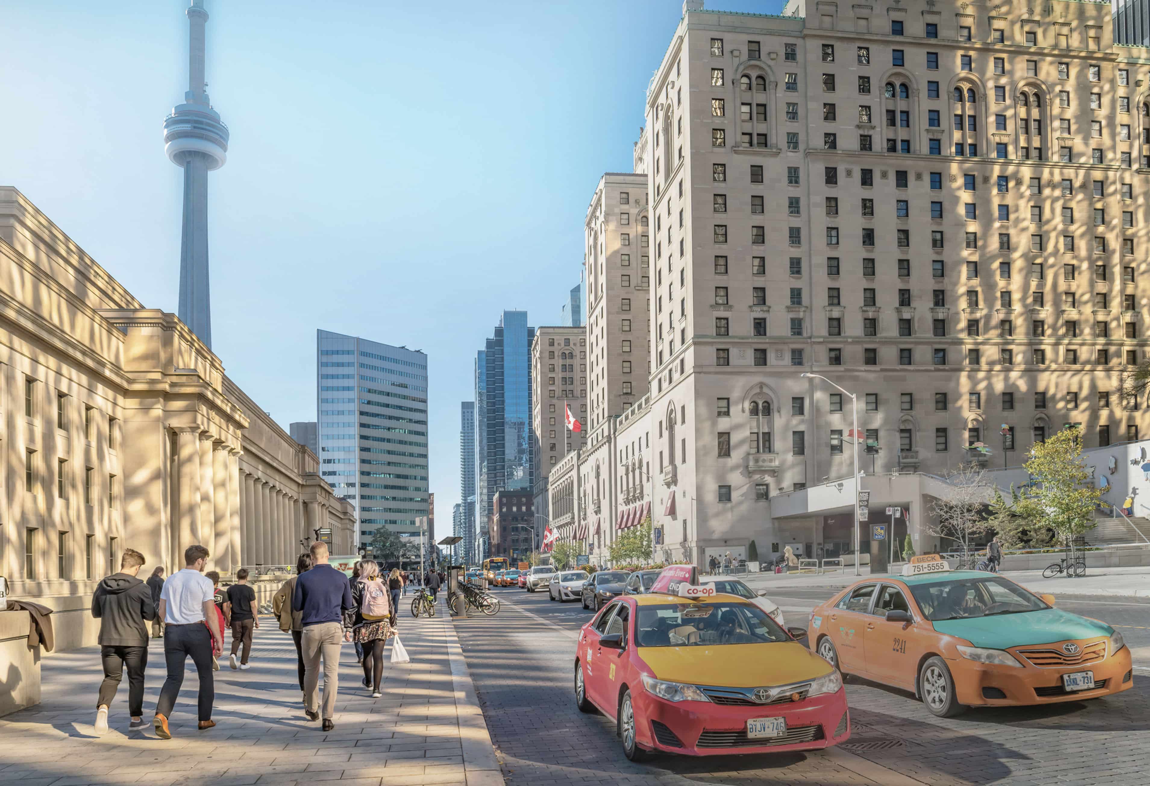 Street scene with pedestrians and taxis near large buildings, including a skyscraper with a needle-shaped top, in an urban landscape under a clear sky.