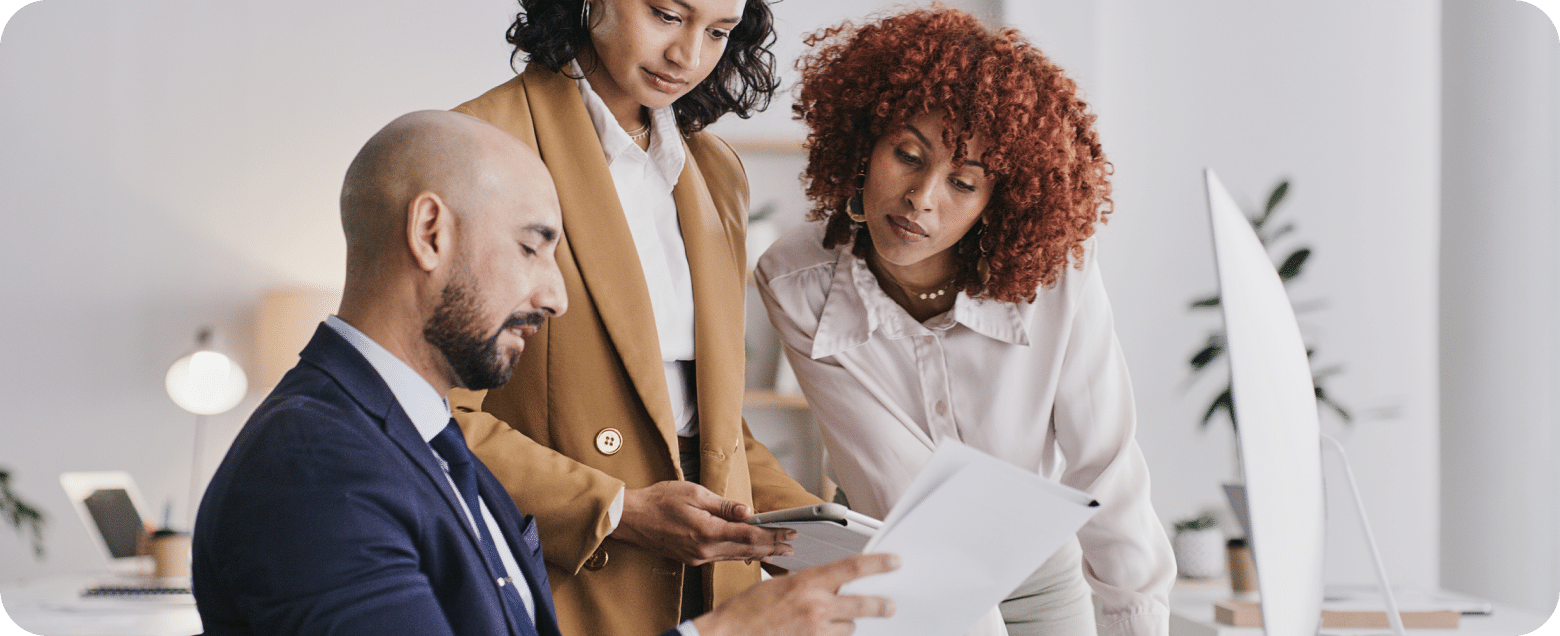 Three people in an office review a document together. One is seated with a paper, while the other two stand close, looking at it attentively.