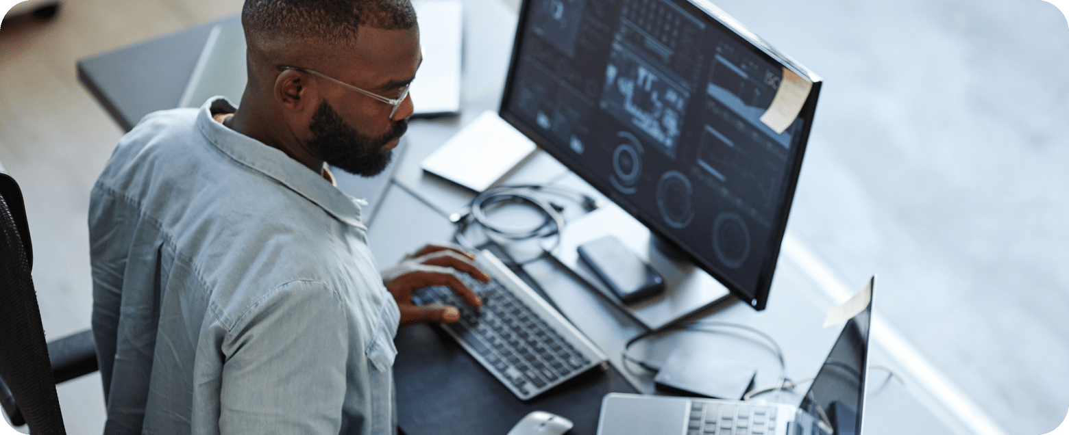 Man working at a desk with two computer monitors displaying data, using a keyboard.