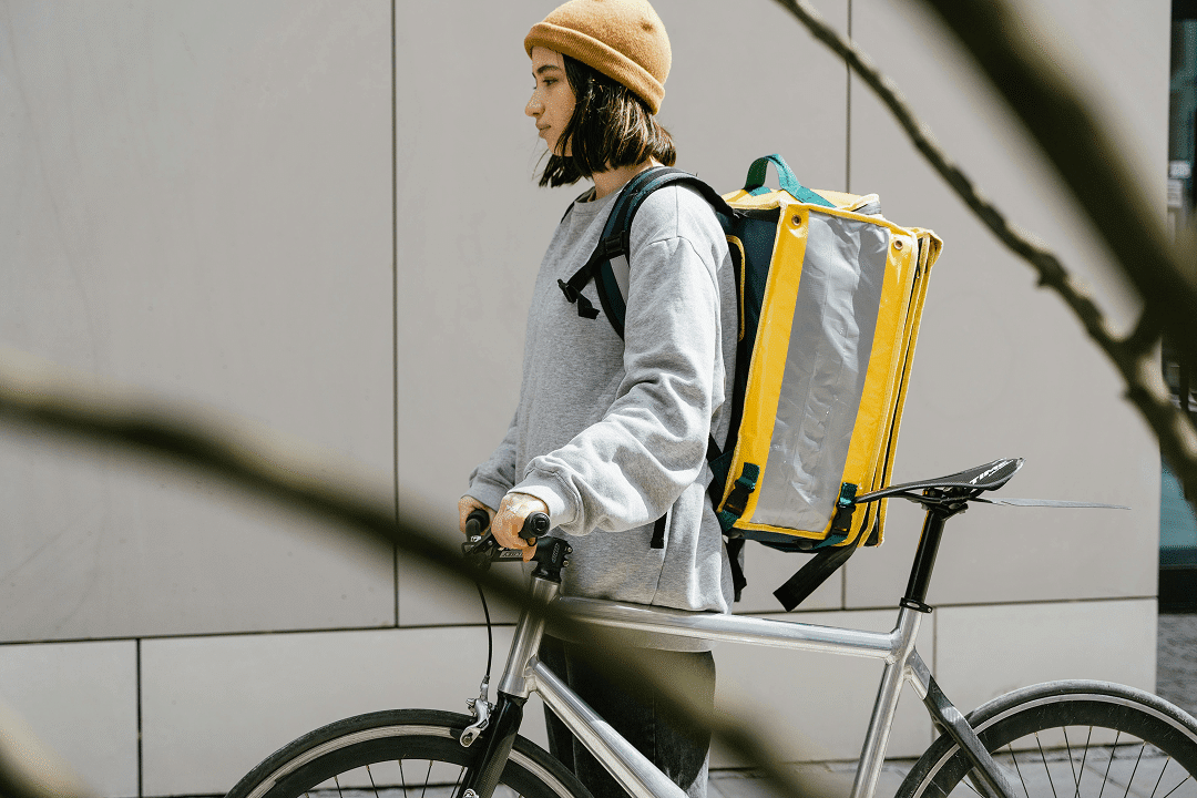 A person wearing a beanie and a large yellow backpack stands next to a bicycle, holding the handlebars, promoting 2025 MP Awareness. A neutral wall serves as the backdrop, with some branches visible in the foreground.