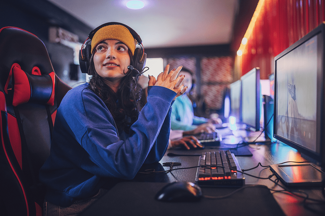 A woman wearing a headset and yellow beanie sits at a computer gaming station, looking to the side. Multiple monitors and players are visible in the background, reflecting the growing MP Awareness leading into 2025.