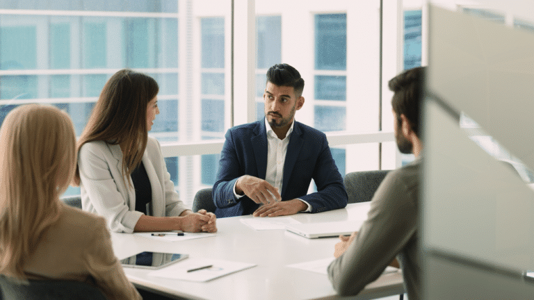 Four people in business attire sit around a conference table in a modern office, engaged in a discussion, with papers and digital devices on the table.