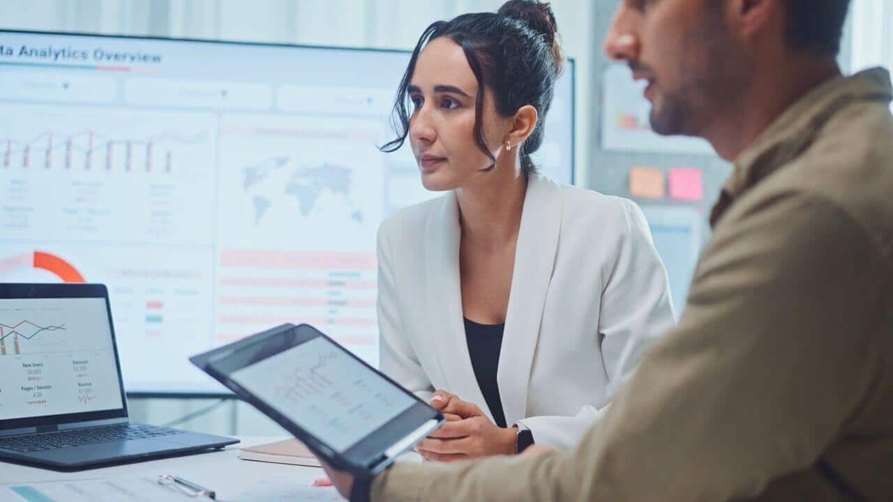 Two professionals discuss data shown on a laptop and tablet in an office, with graphs and charts displayed on a screen in the background.