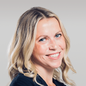 A woman with long blonde hair and blue eyes smiles at the camera against a plain, light gray background.