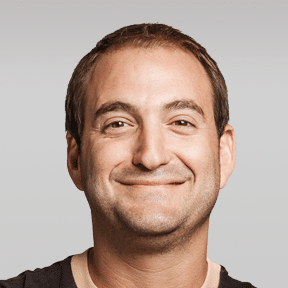 A man with short brown hair is smiling at the camera against a plain light gray background.