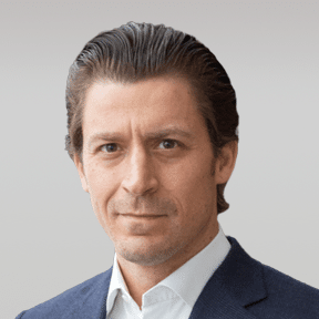 A man with slicked-back brown hair wearing a dark blue suit jacket and white collared shirt, facing forward against a plain light background.