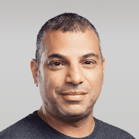 A man with short, dark hair and a trimmed beard, wearing a dark t-shirt, poses against a plain, light gray background.
