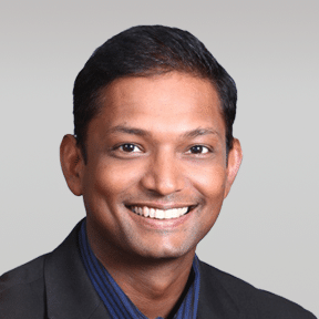 A man with short dark hair is smiling, wearing a dark blazer and a blue collared shirt, posed against a plain light grey background.