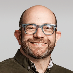 A man with a beard and glasses smiles at the camera against a plain, light gray background.