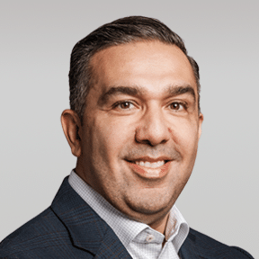 Man with short dark hair wearing a checked suit jacket and white dress shirt, smiling at the camera against a plain light gray background.