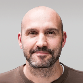 A middle-aged man with a bald head, trimmed beard, and mustache, wearing a brown crew-neck shirt, smiles at the camera against a plain gray background.