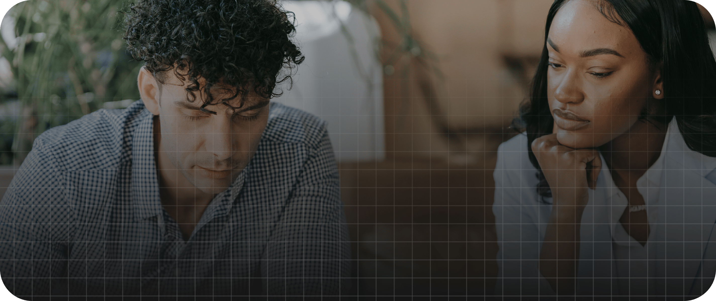 A man and a woman sit next to each other at a table, both looking down with serious expressions.