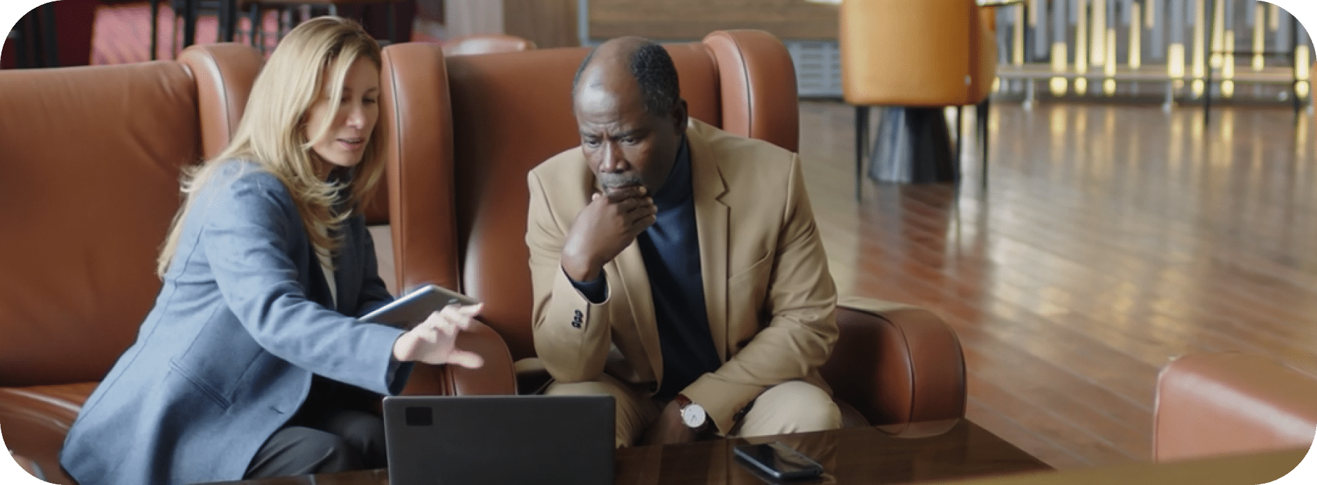Two business professionals sit in armchairs, looking intently at a laptop screen on a coffee table in a modern office setting.