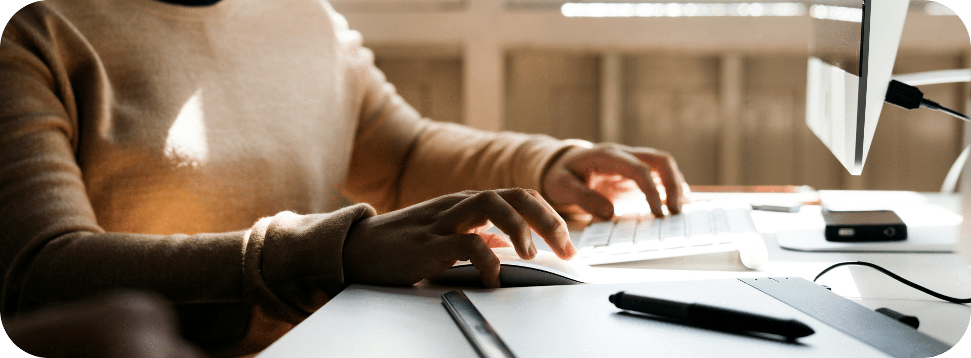 Person typing on a computer keyboard at a desk with a pen and tablet in the foreground, in a well-lit room.