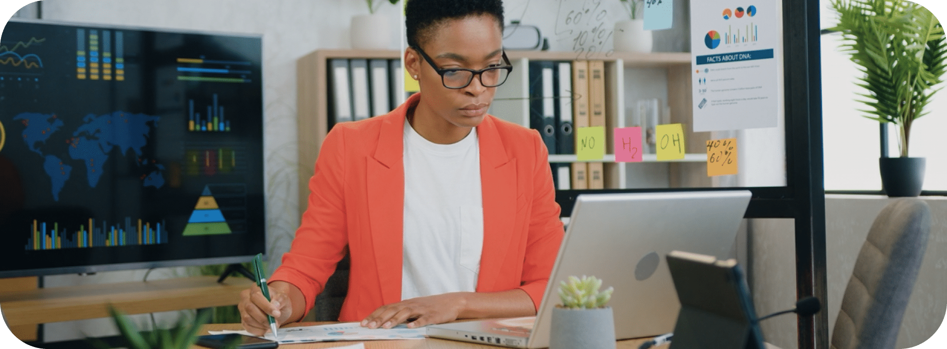 Person in a red blazer working at a desk with a laptop, taking notes, and analyzing charts in a modern office setting with plants and graphs in the background.