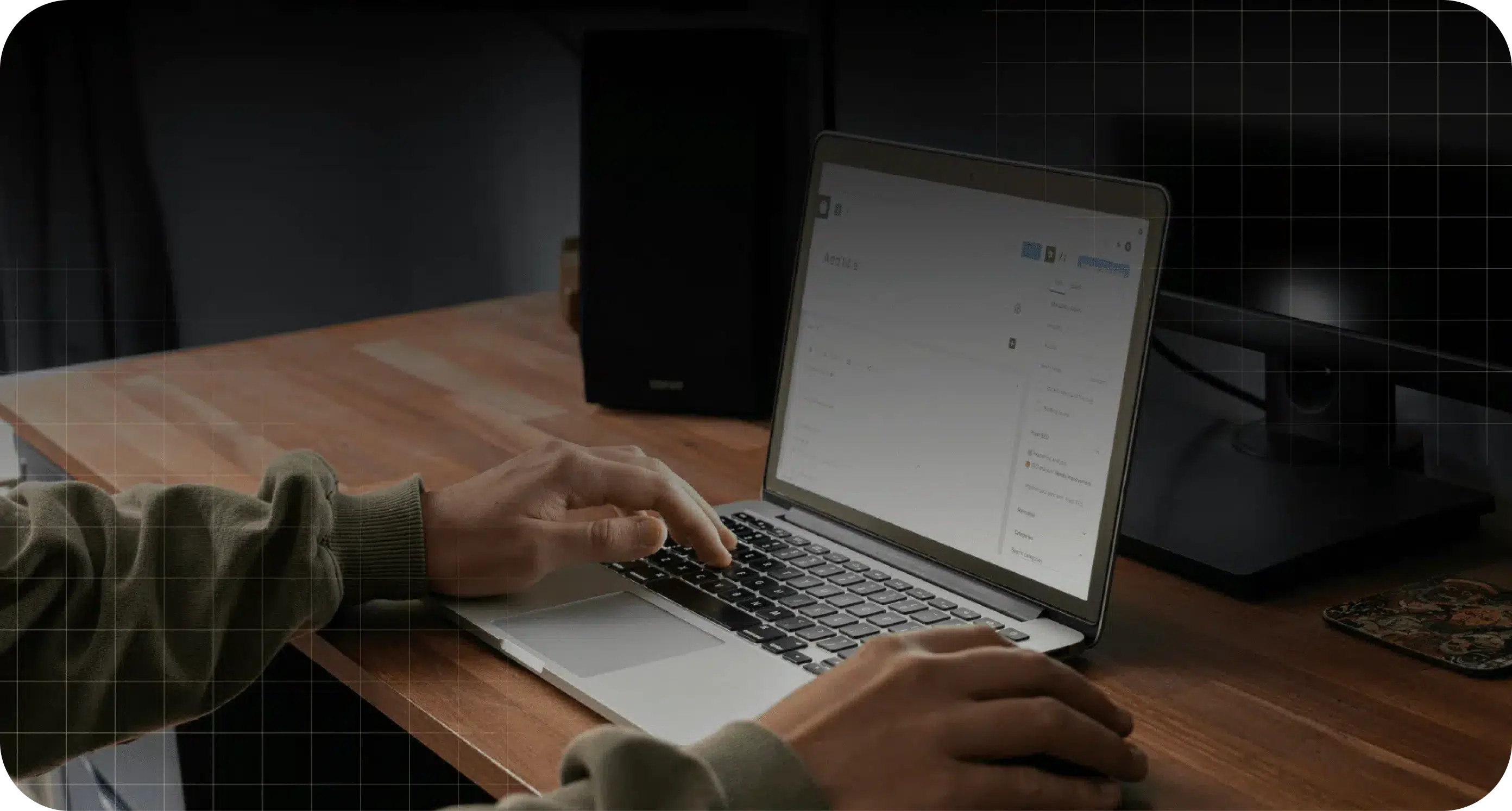 Person typing on a laptop at a wooden desk with a monitor, mousepad, and speaker in the background.